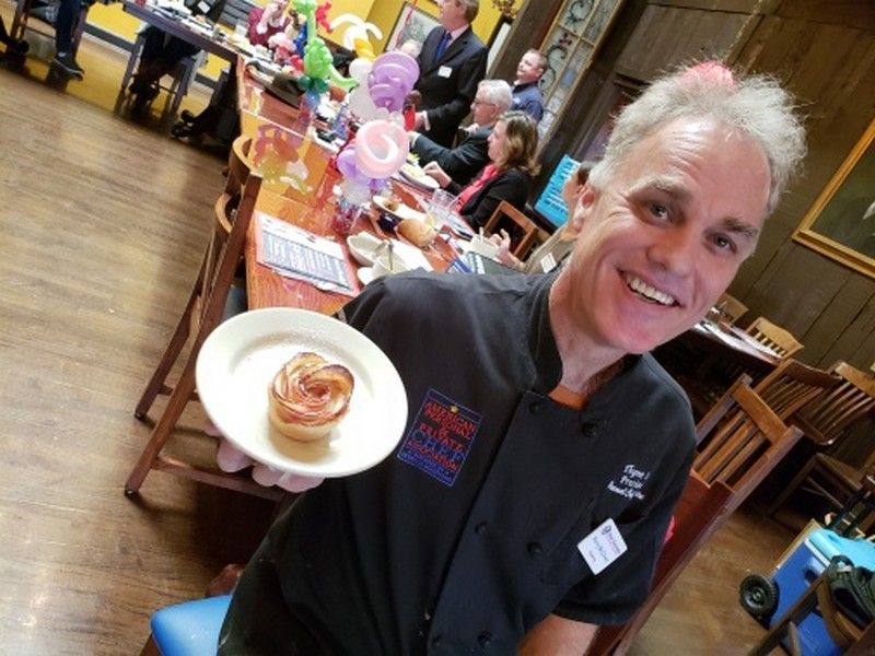 A chef in black uniform holding up a plate with a pastry, smiling at the camera, with seated guests and balloons in the background