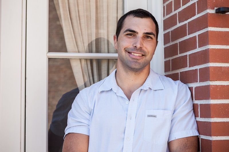 Man in blue checkered shirt smiling in front of a house window and brick wall